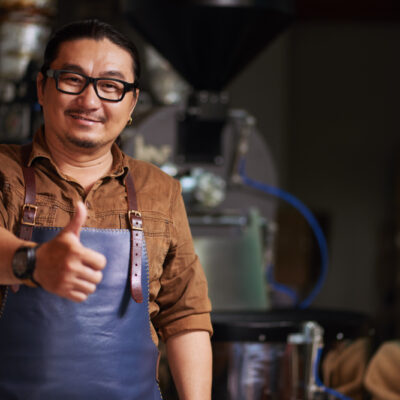 middle-aged-asian-man-posing-with-thumb-up-front-coffee-roasting-equipment